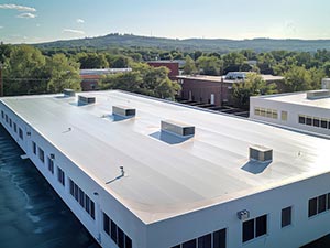 Single-Ply-Roofing-Butte-MT-Montana-2 | Schrock Roofing Aerial view of a large white flat-roofed commercial building with multiple skylights and vents, surrounded by trees and distant hills in blue sky.