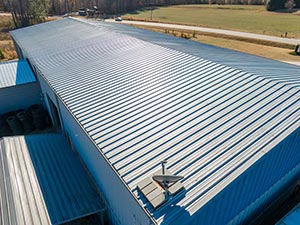Standing-Seam-Metal-Roof-Missoula-MT-Montana-1 | Schrock Roofing Blue corrugated metal roof of a low industrial building with a security camera along the edge; a parking lot and grassy field are in the background.